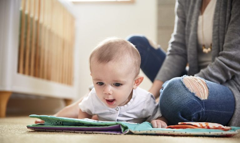 A baby practices tummy time on a playmat with their mother offering support in the background.