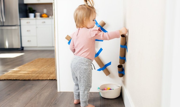 Toddler putting a ball through paper towel rolls that are taped to the wall