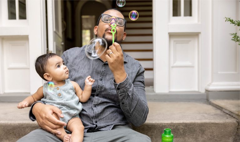 Dad blows bubbles with child on his lap.