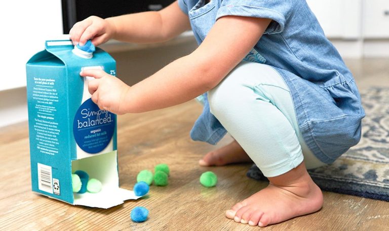 Toddler putting pom poms in an empty cardboard container